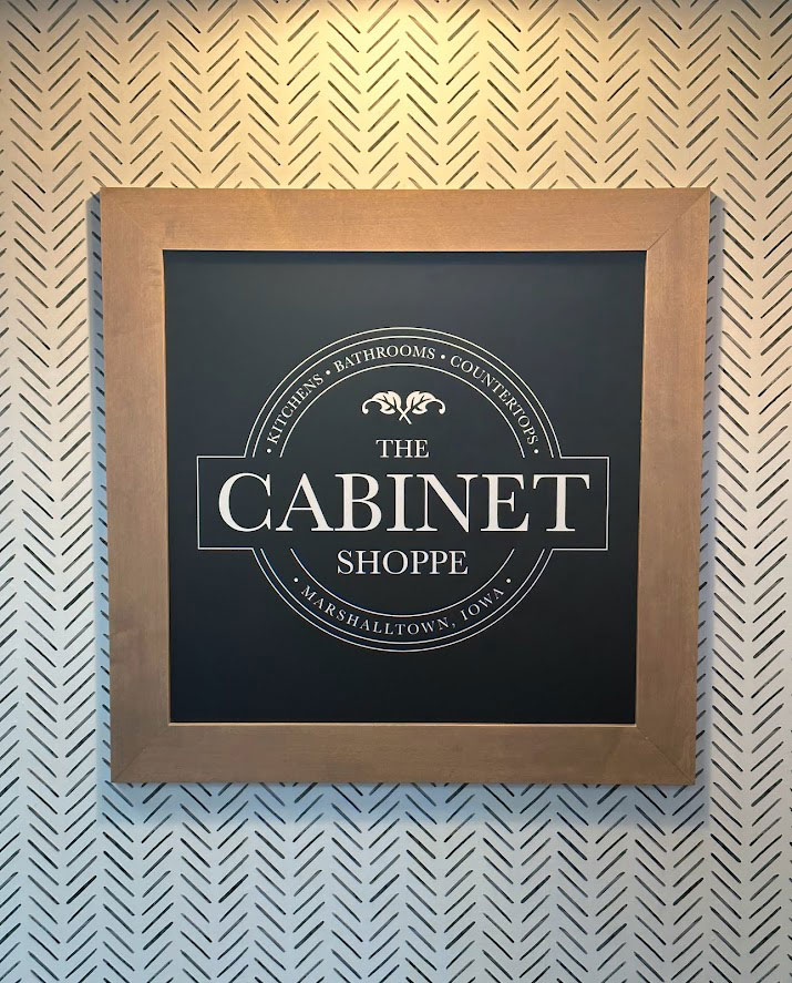 Framed picture of The Cabinet Shoppe logo hung up on white and black herringbone patterned wallpaper.