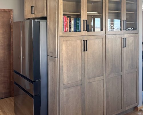 Kitchen remodel with medium brown cabinets wrapping around the corner from a fridge. The cabinets are floor-to-ceiling and the top cabinets have glass doors.